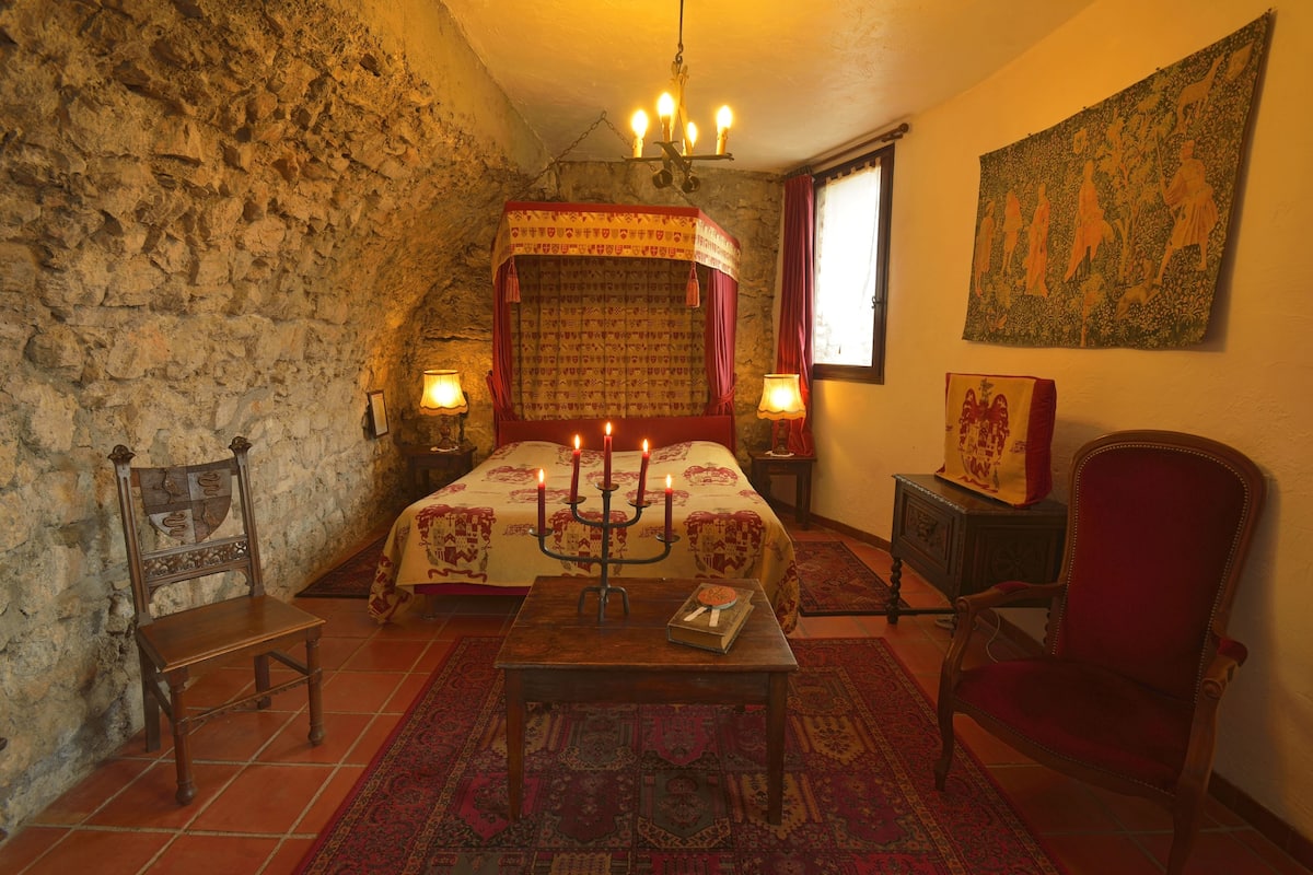 A traditional room with a stone wall, a bed with a canopy, a candelabra, a chair, and a tapestry.