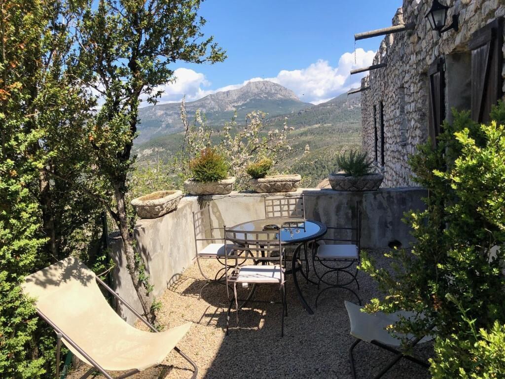 Une terrasse extérieure avec une table et des chaises, entourée de murs en pierre et de plantes en pot, offrant une vue sur un paysage montagneux.