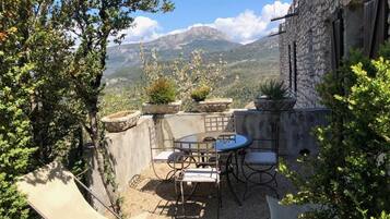 Une terrasse extérieure avec une table et des chaises, entourée de murs en pierre et de plantes en pot, offrant une vue sur un paysage montagneux.