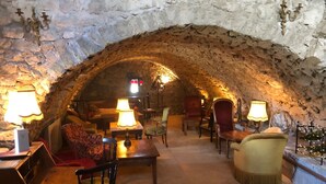 Una habitación acogedora con paredes de piedra y techo abovedado, amueblada con un sofá, sillas y mesas, iluminada por lámparas.