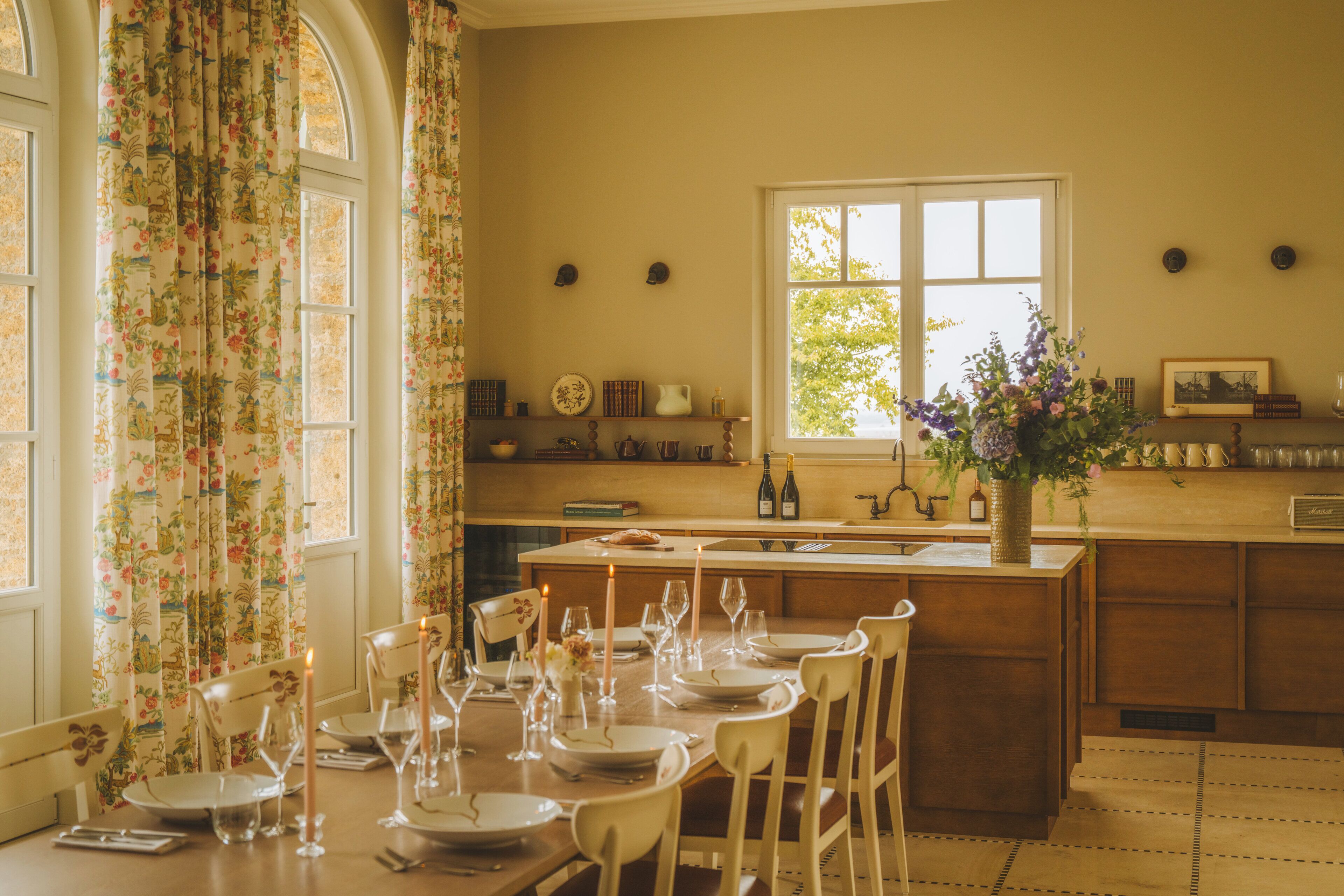 Un coin repas avec une table en bois dressée pour quatre personnes, entourée de chaises blanches. Un vase rempli de fleurs est posé sur la table, et des bouteilles de vin et des verres se trouvent sur le comptoir.