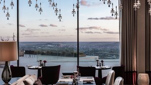 Un área de comedor con una mesa dispuesta para dos, vista panorámica de la ciudad a través de amplias ventanas y candelabros colgantes.