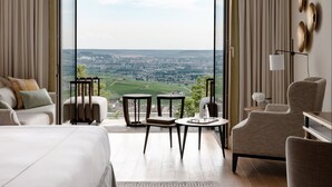 Habitación de hotel moderna con una cama grande, dos mesitas de noche, un sofá y vistas a un paisaje urbano a través de ventanas de gran altura.