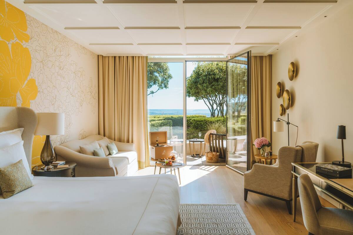 Une chambre d’hôtel moderne avec un grand lit, un coin salon comprenant un canapé et des fauteuils, une petite table avec un vase et un livre, et une vue sur l’extérieur grâce à de grandes portes vitrées.