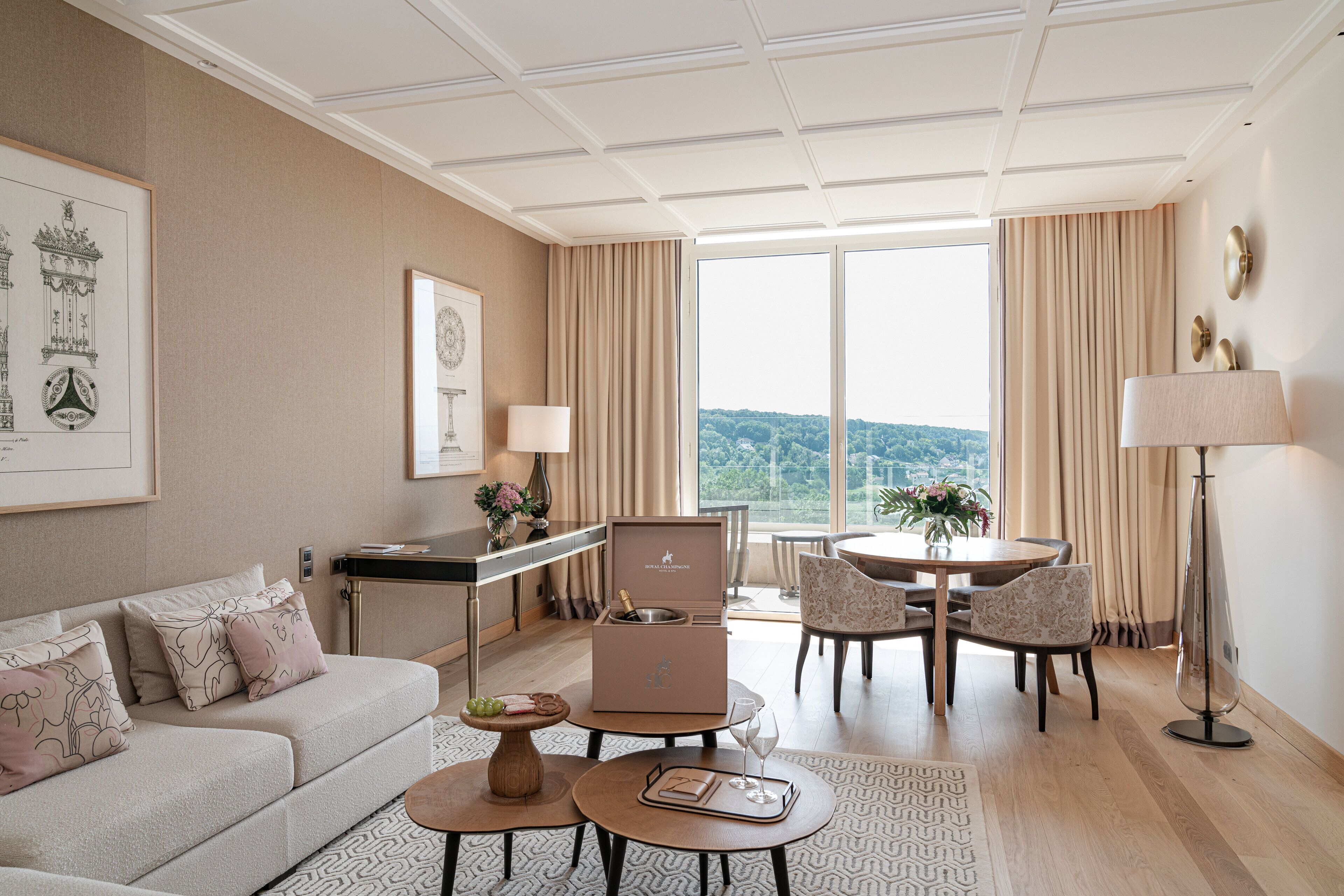 A modern hotel room with a sofa, dining table, and chairs, a minibar, and a large window with a view.