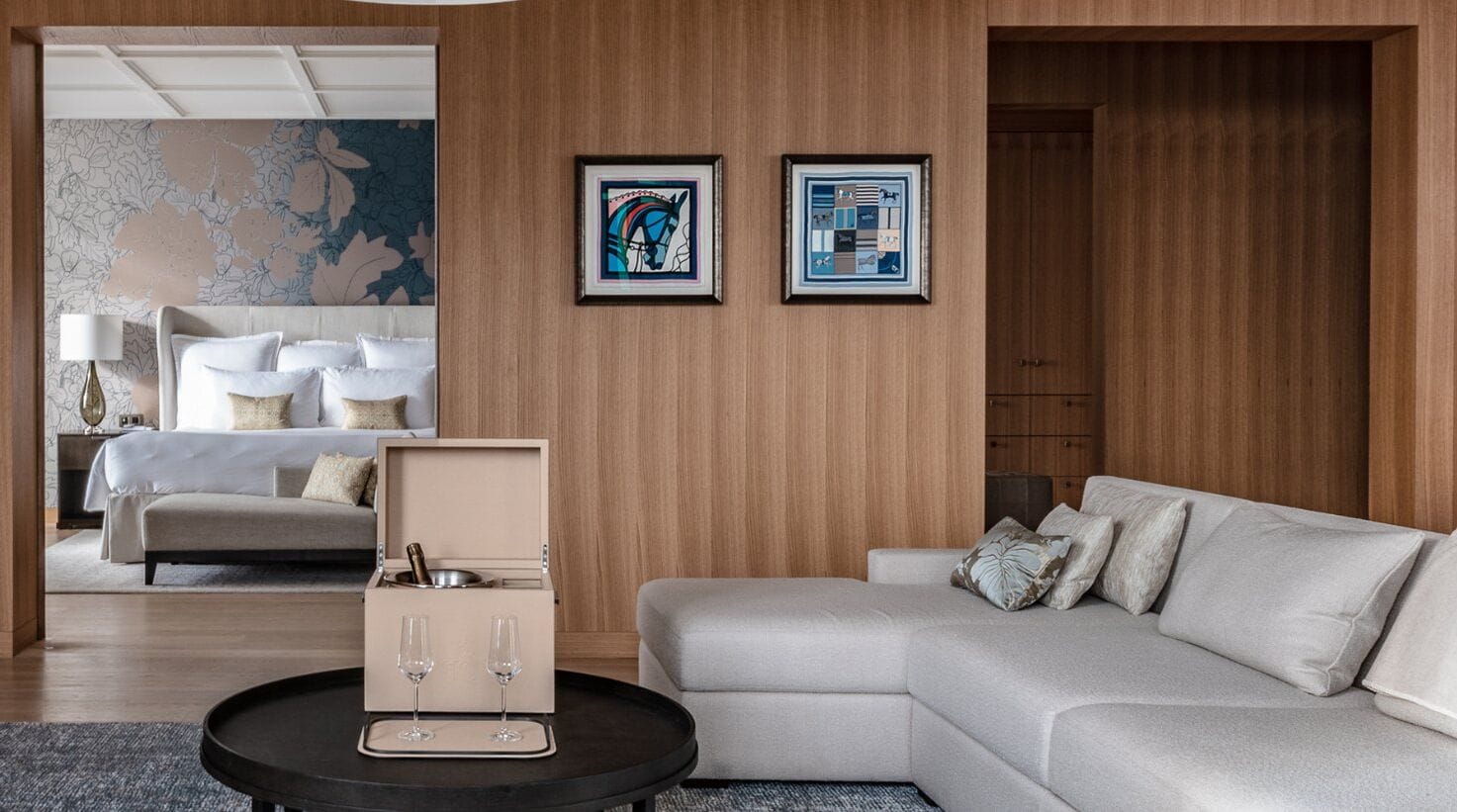 A modern hotel room with a beige sofa, a black coffee table, and a bed with white linens. There are two framed artworks on the wall and a wooden wardrobe.