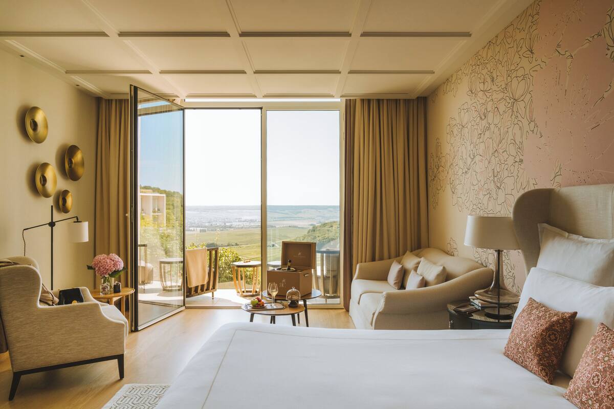 Une chambre d’hôtel avec un grand lit, un canapé, un fauteuil, une petite table avec une lampe et une vue sur l’extérieur.