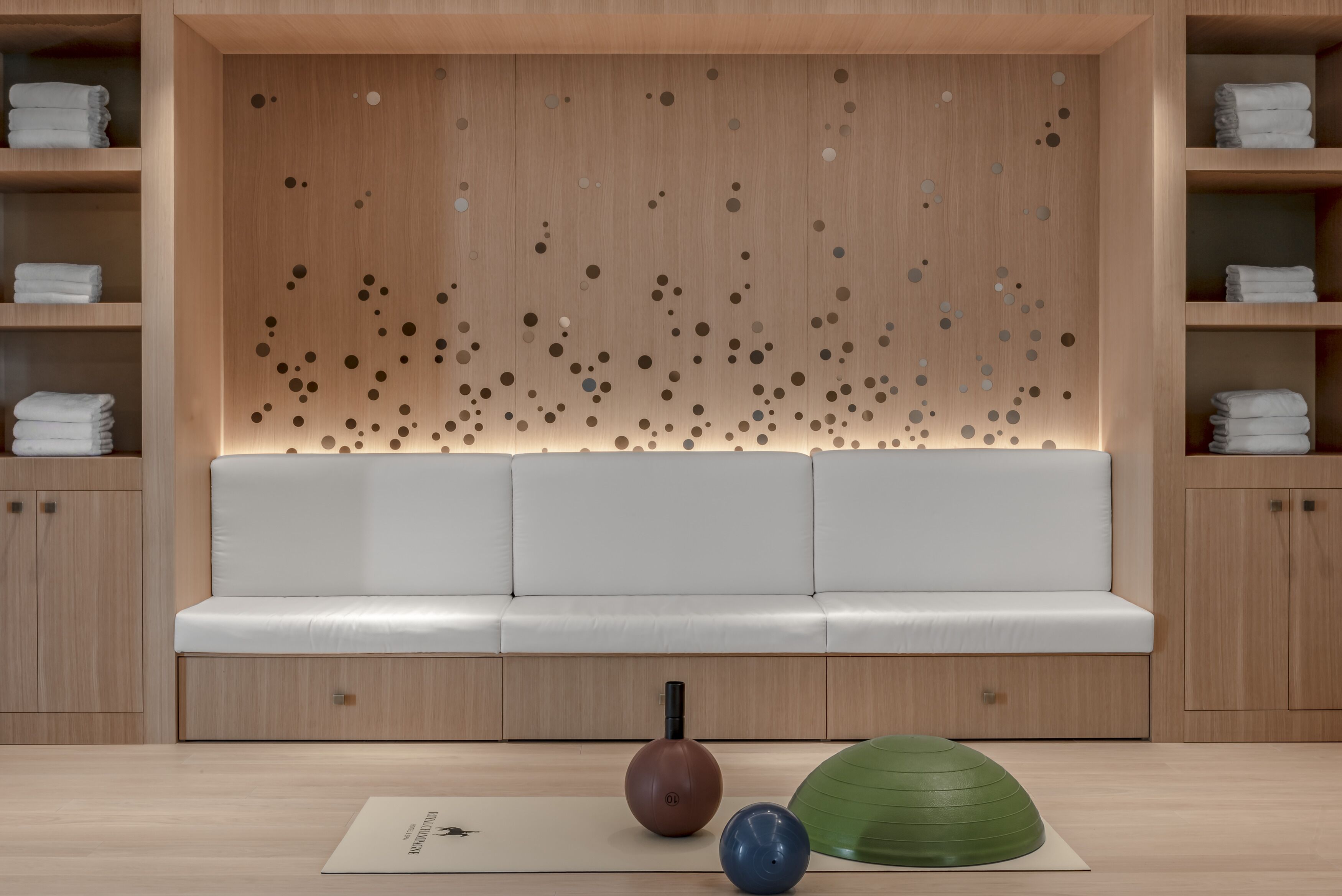 A modern, minimalist room with a wooden wall featuring circular patterns, a white sofa, and neatly arranged towels on shelves.