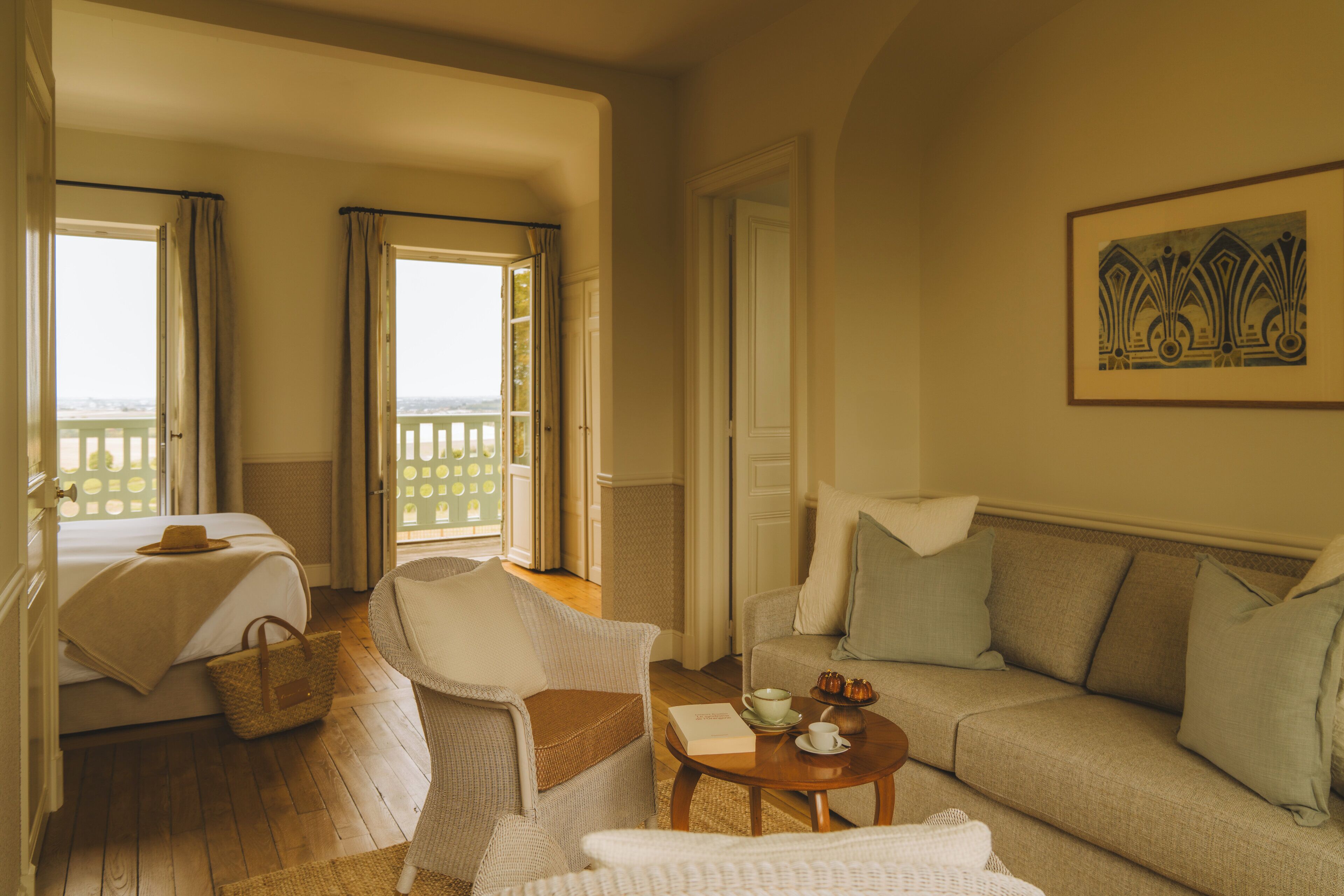 A room with a bed, two chairs, a sofa, a table with a book and a cup, and a view through the window.