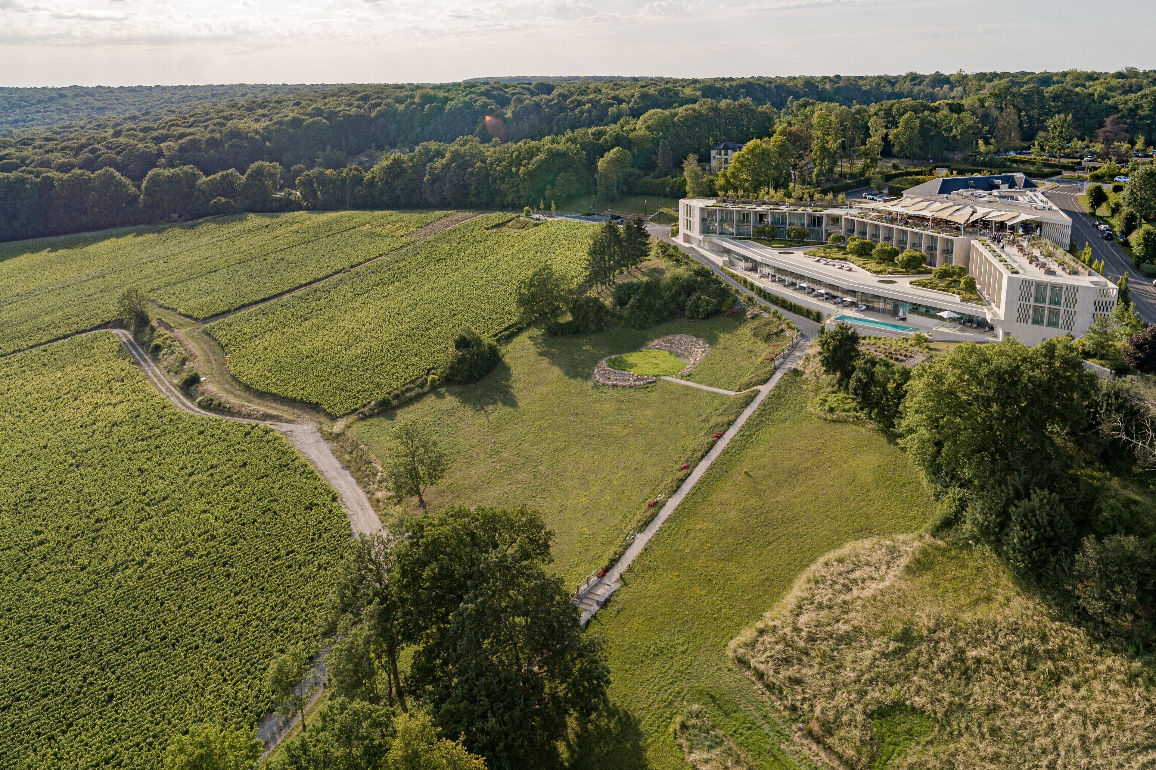 An aerial view of a modern building with a large glass facade, surrounded by lush green fields and a small pond.