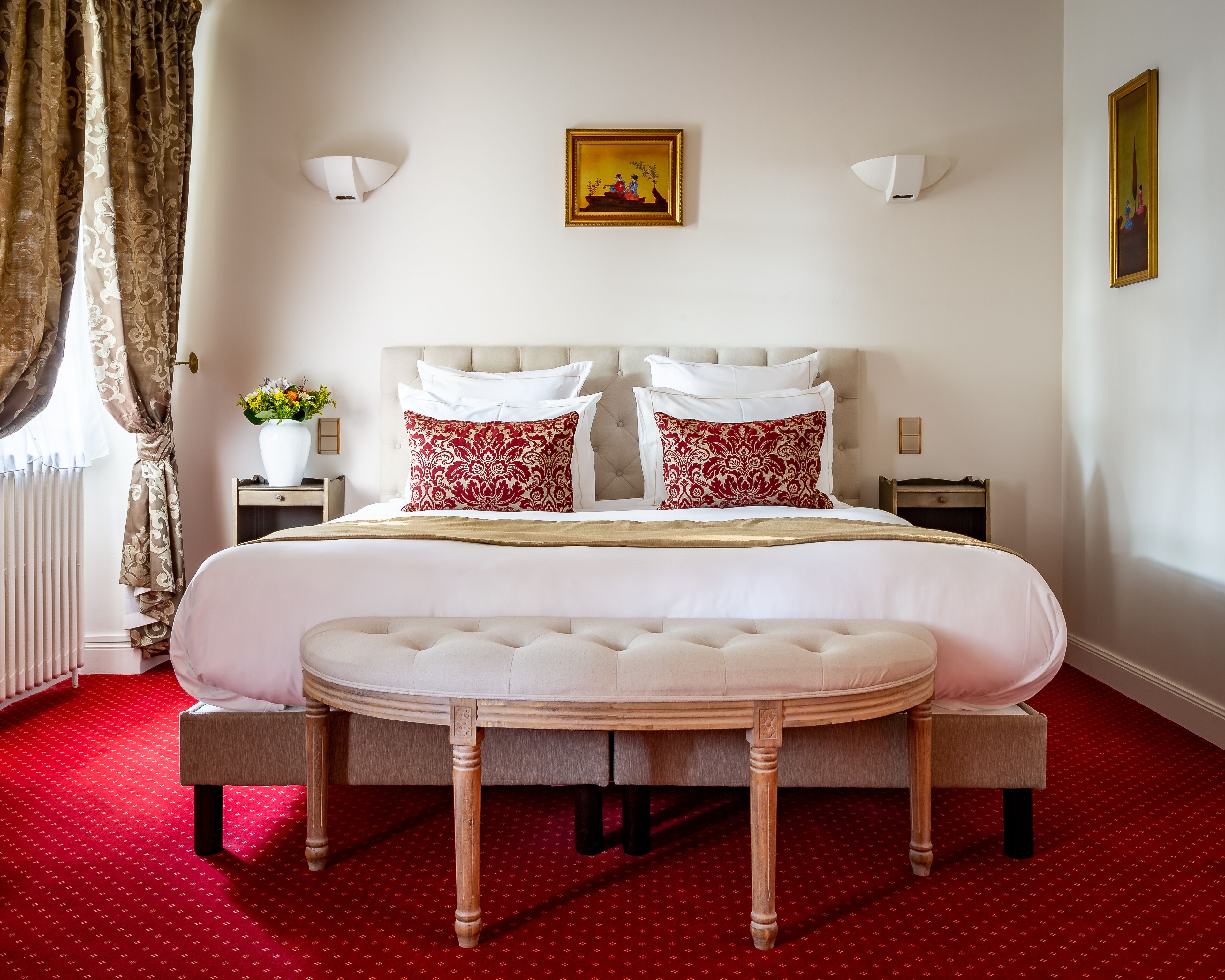 Un lit bien fait, avec des oreillers blancs et rouges, une table de chevet ornée d’un vase de fleurs et une lampe fixée au mur.