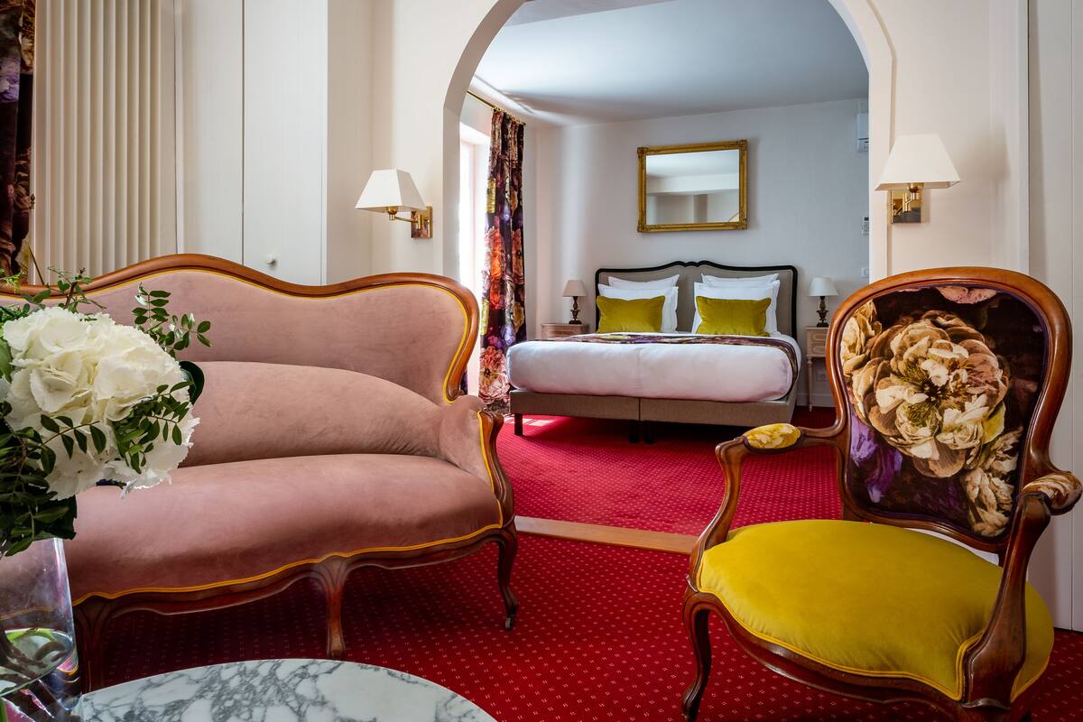 Habitación de hotel con cama, dos sillones, alfombra roja y un cuadro floral en la pared.