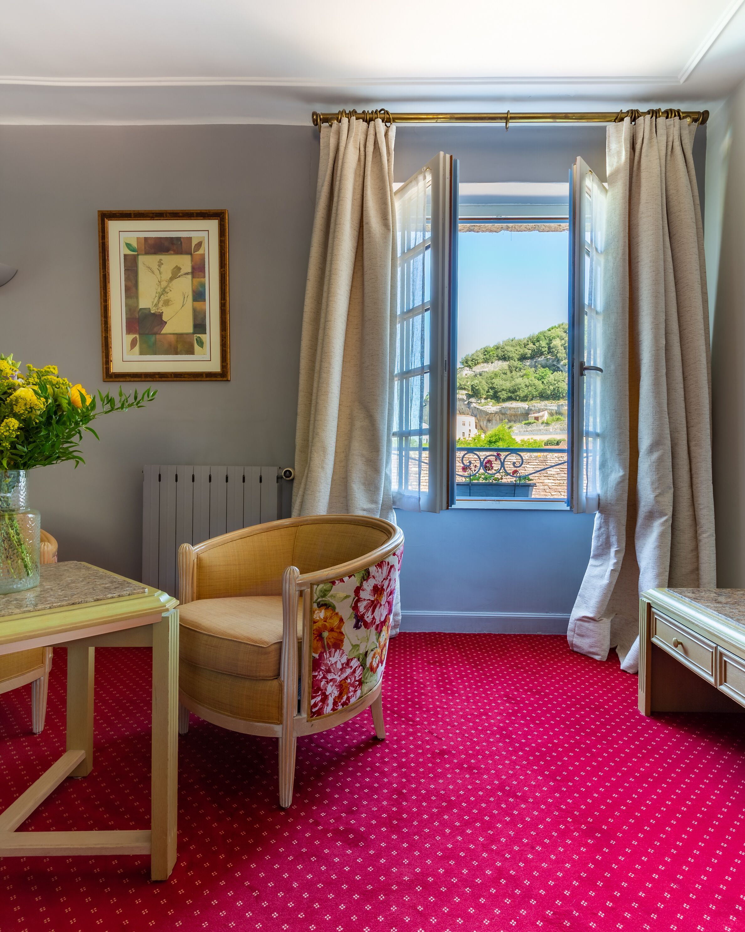 A room with a red patterned carpet, a wooden chair with floral upholstery, a table with a vase of flowers, a framed picture on the wall, and a window with curtains.