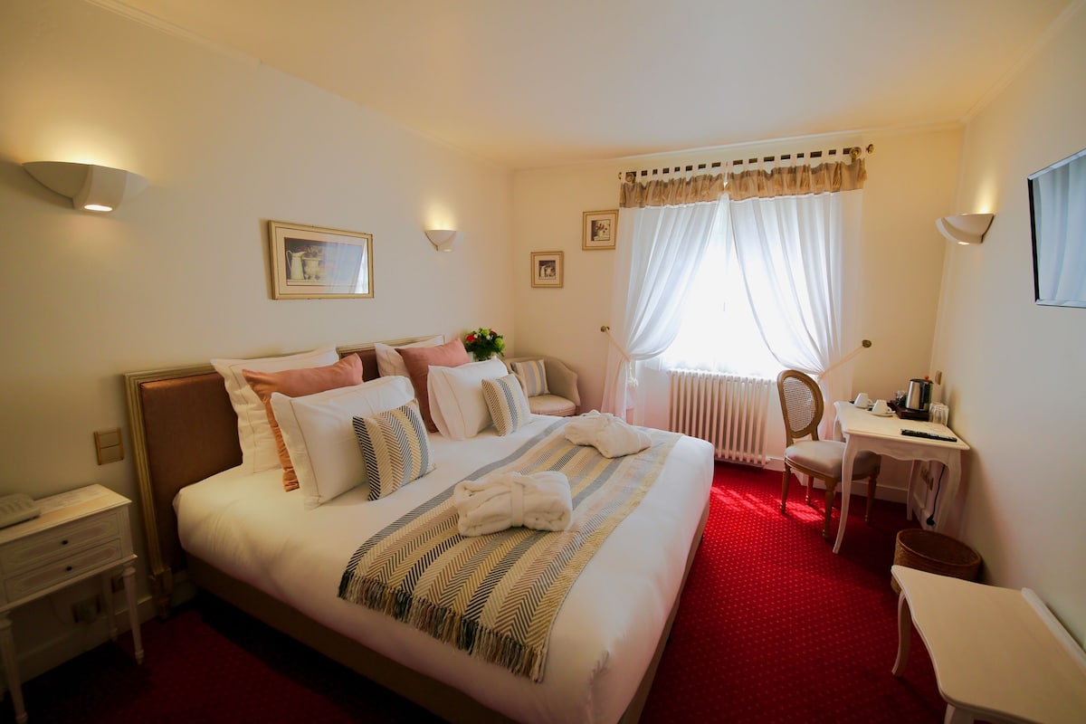 Habitación de hotel con una cama grande, un escritorio y una silla. Cuenta con lámparas de pared y un ventanal con cortinas.