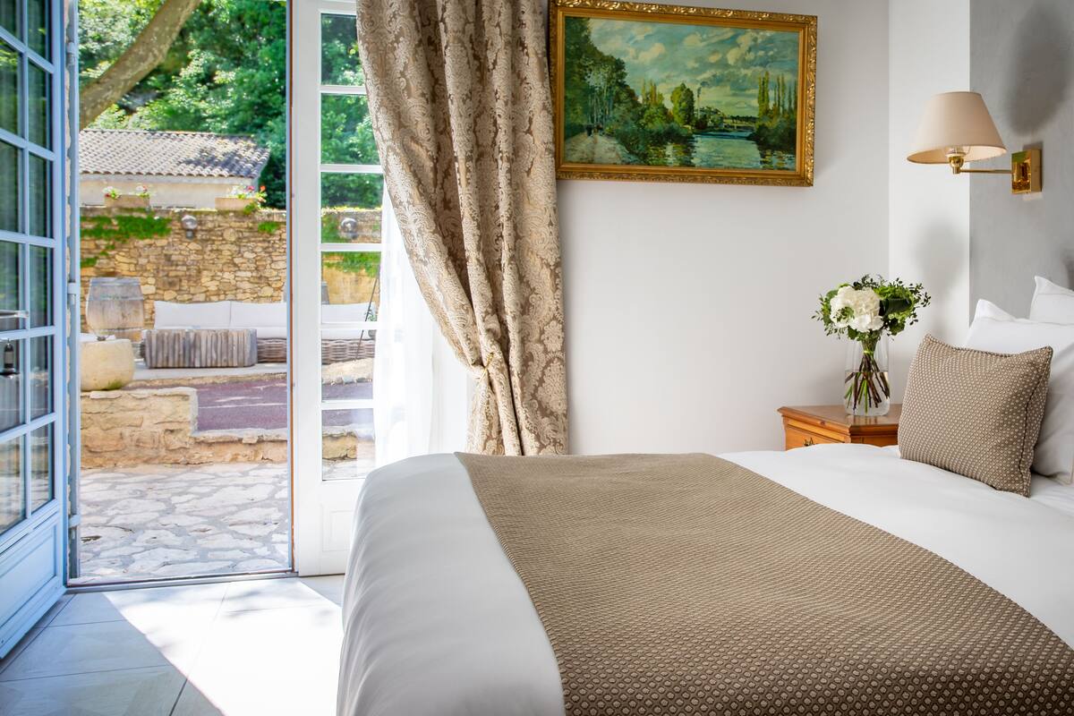 Un dormitorio con una cama grande, un cuadro en la pared, una mesita de noche con una lámpara y vistas a un jardín a través de una puerta abierta.