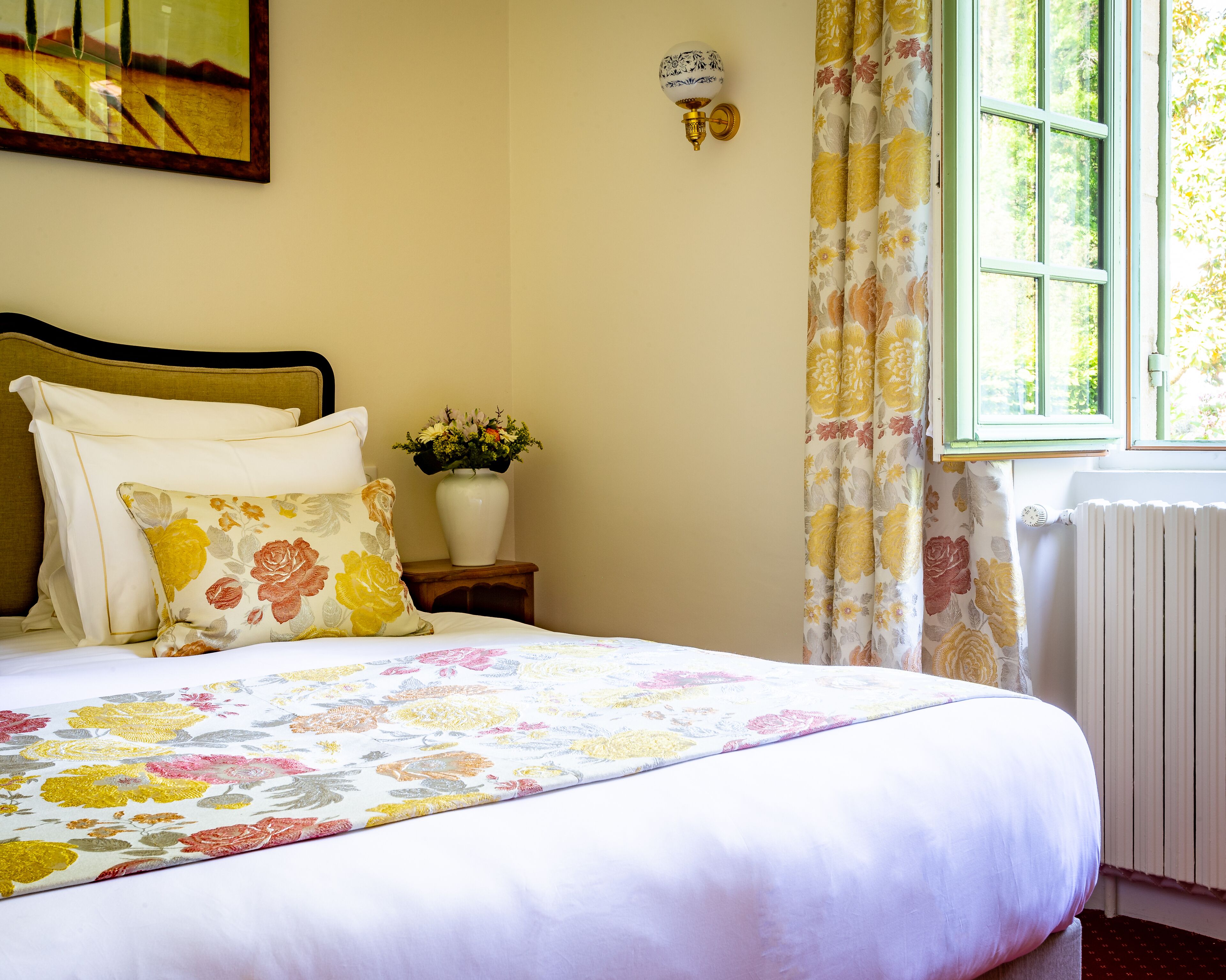 A neatly made bed with floral bedding, a vase of flowers on a bedside table, and a window with curtains.