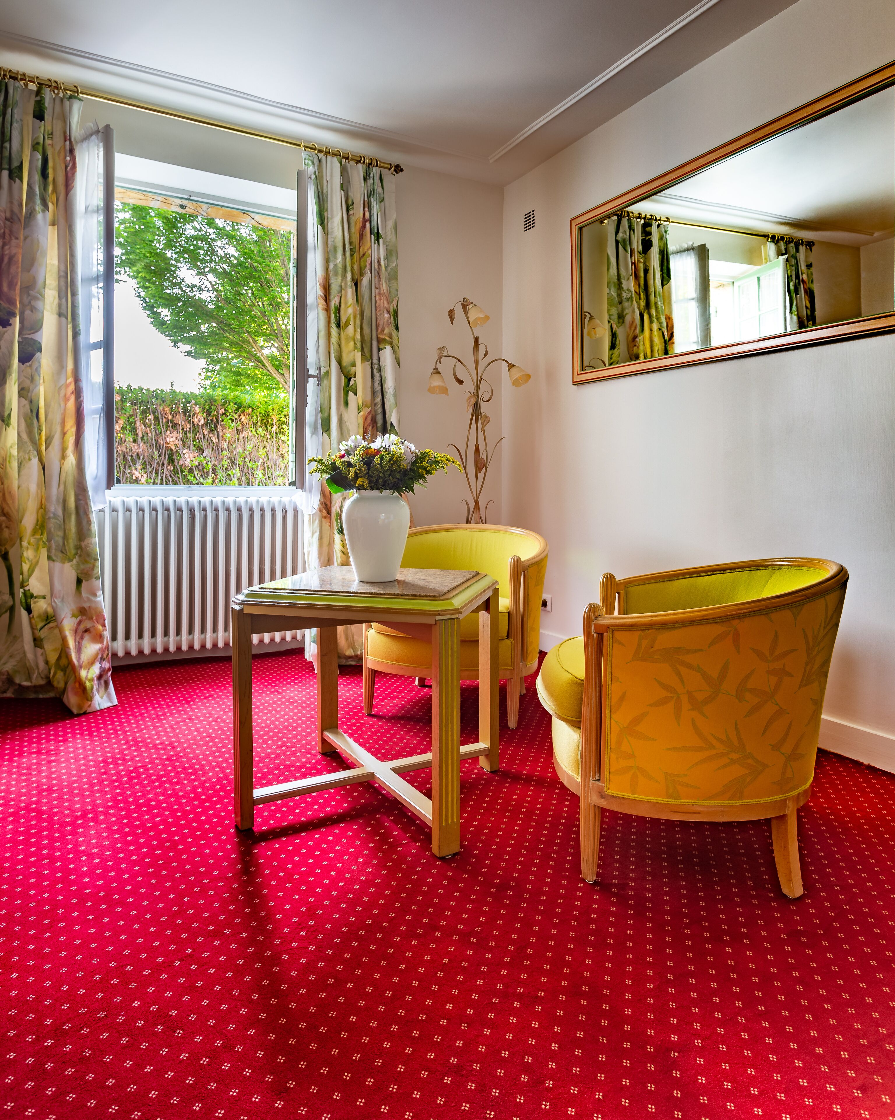Une pièce avec un tapis rouge à motifs, une table sur laquelle est posé un vase de fleurs, deux chaises jaunes et un grand miroir au mur.