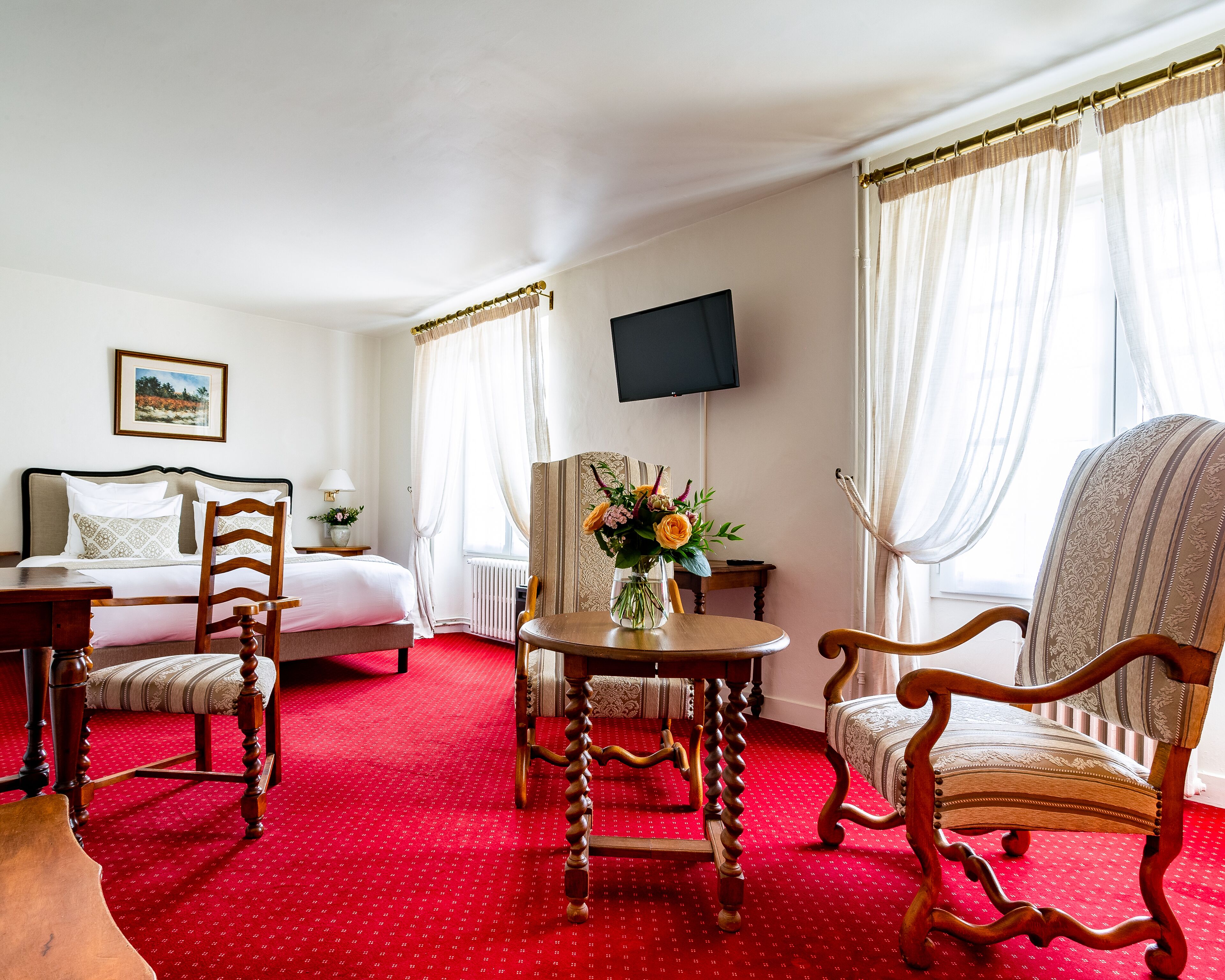 Habitación de hotel con una cama, dos sillas de madera, una mesita con un florero y una televisión montada en la pared, además de una alfombra roja.