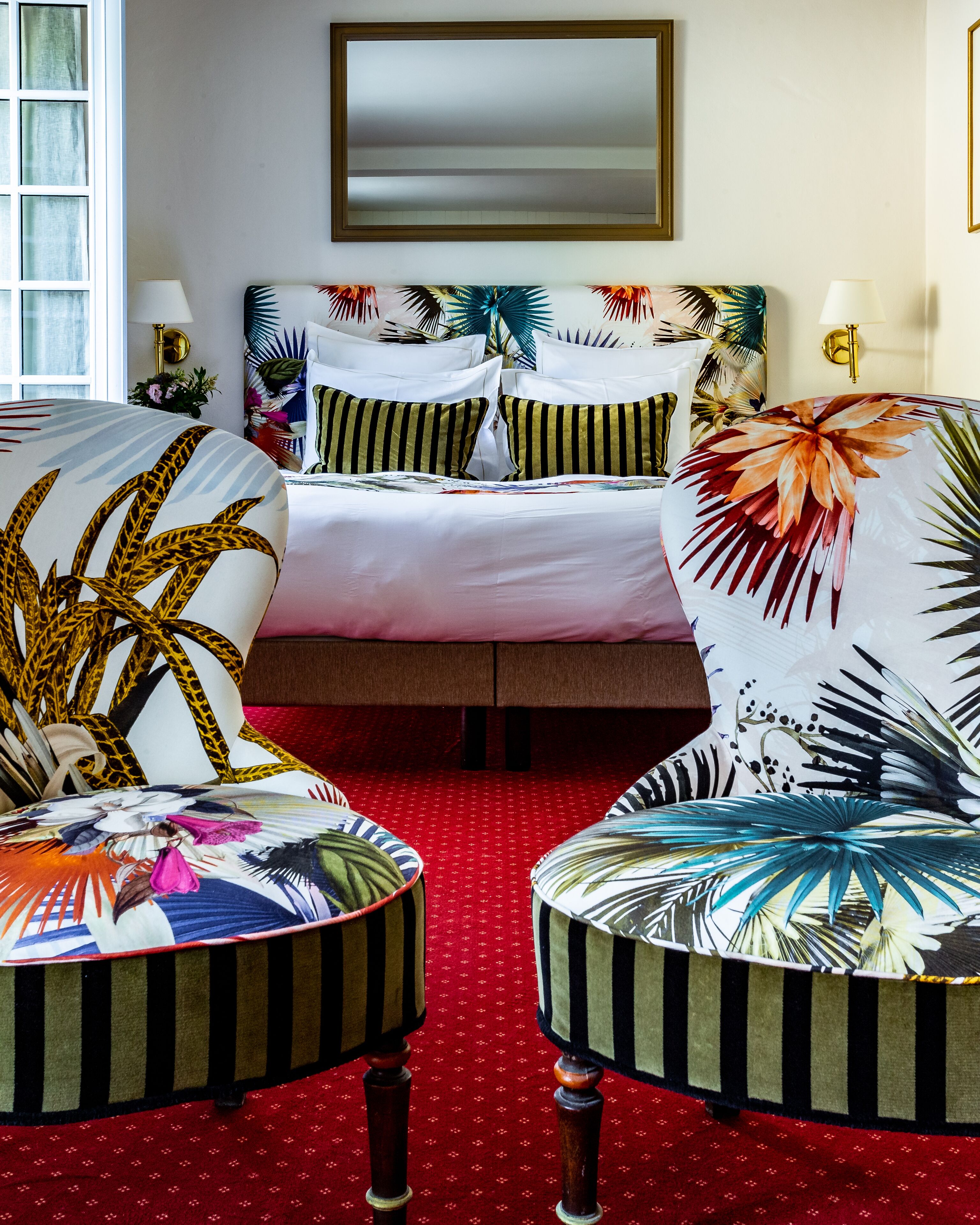 Une chambre d’hôtel avec un lit, deux fauteuils aux motifs tropicaux, une moquette rouge et un grand miroir au mur.