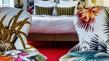 Une chambre d’hôtel avec un lit, deux fauteuils aux motifs tropicaux, une moquette rouge et un grand miroir au mur.