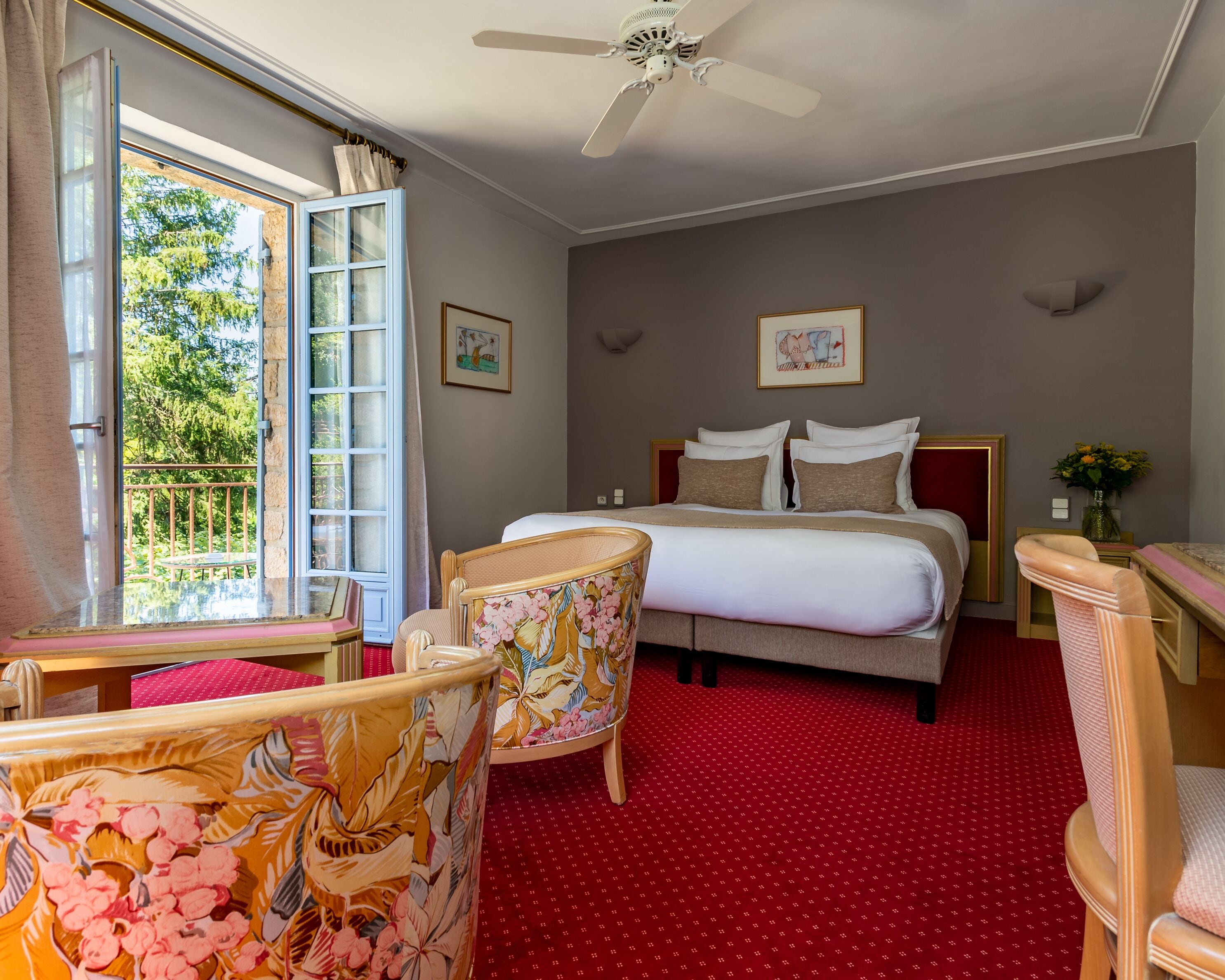 A bedroom with a bed, two chairs, a ceiling fan, and a window with a view of trees.