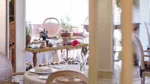 Un comedor con una mesa redonda dispuesta para dos personas, rodeada de sillas de mimbre. Se aprecia una estantería con frascos y una flor al fondo.