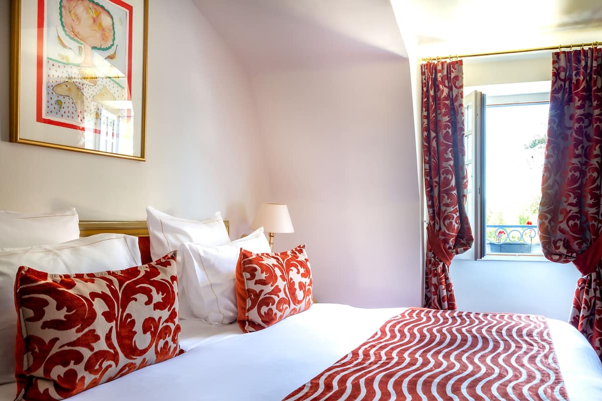 Una habitación de hotel con una cama, almohadillas blancas y rojas con estampado, una colcha blanca y roja a rayas, un cuadro en la pared y una ventana con cortinas.
