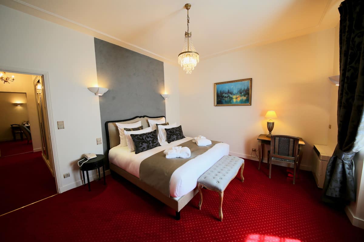 Una habitación de hotel con una cama grande, un escritorio pequeño, una silla, un banco, un candelabro, un cuadro y una alfombra roja.