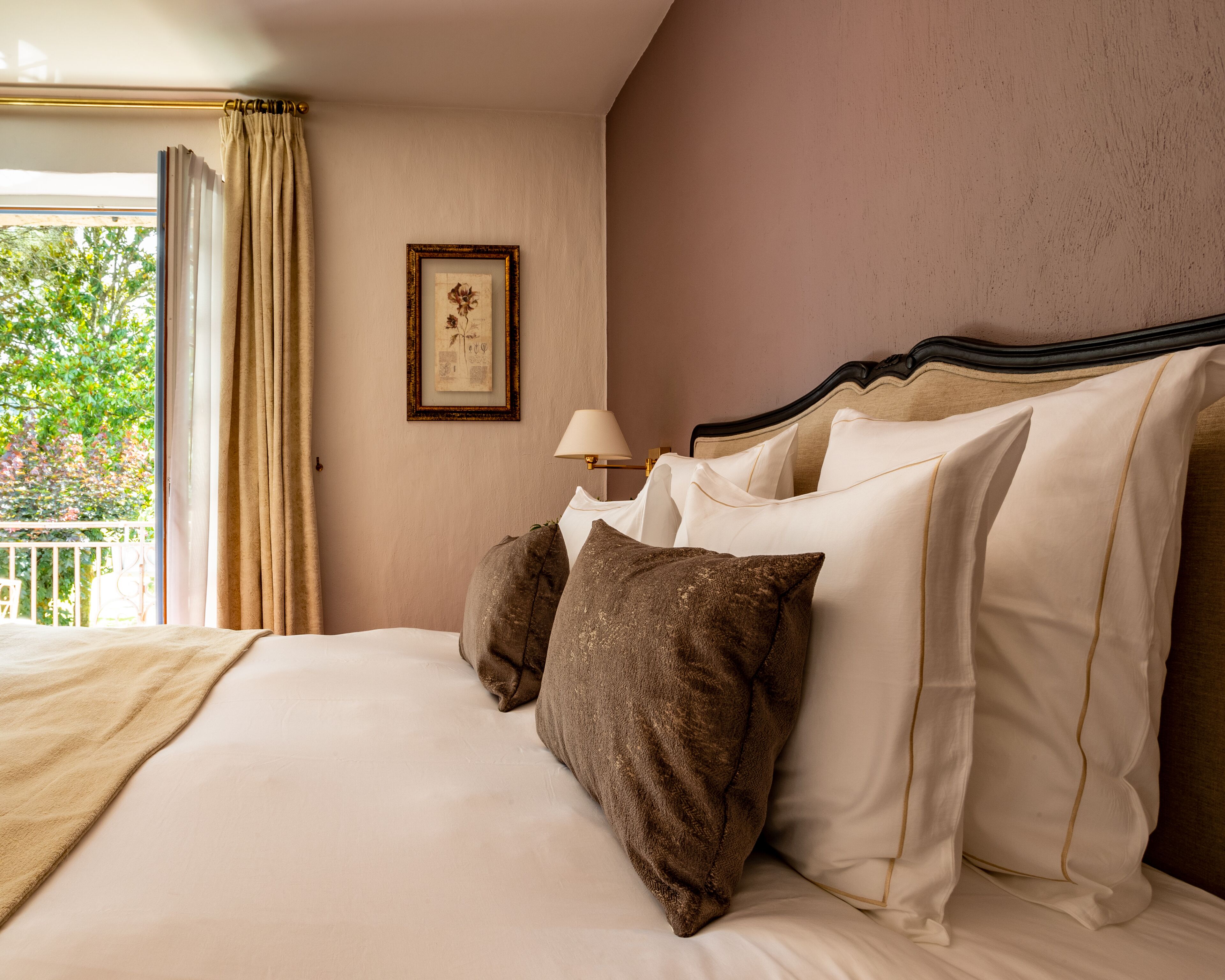 A hotel room with a bed, pillows, a lamp, a framed picture on the wall, and a view of greenery outside.