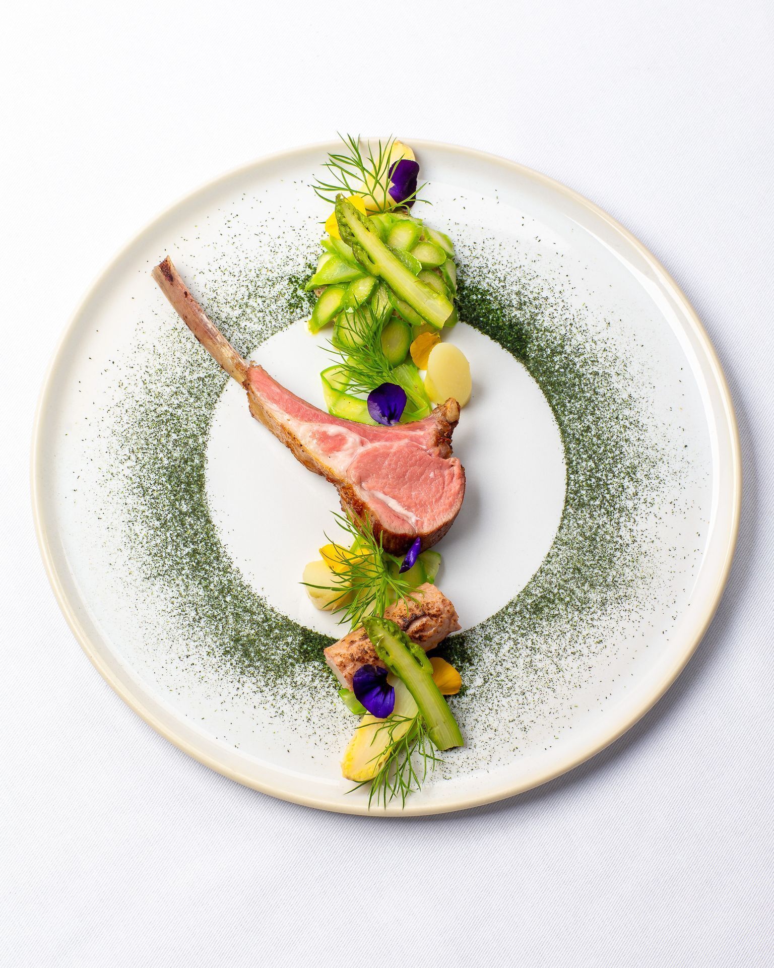 Une assiette contenant une côte d’agneau, des asperges et des pommes de terre, le tout agrémenté d’herbes et de fleurs.