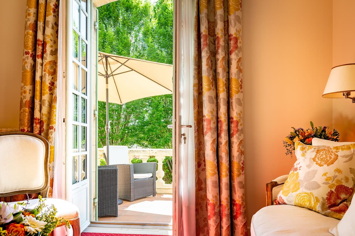 Una habitación con cama, una silla, una mesa con flores y vistas a un patio exterior con mesa y sillas.