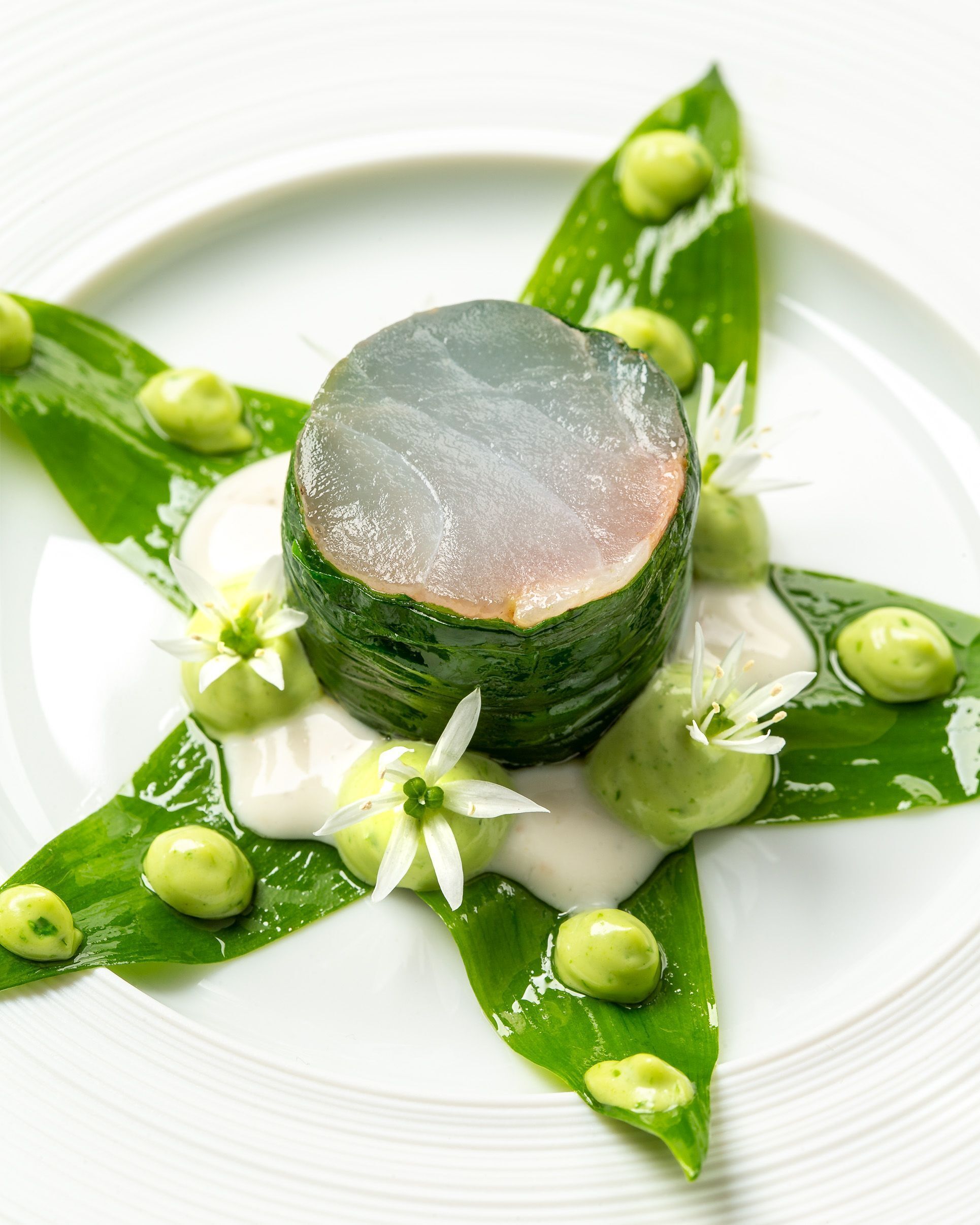 A dish with a central piece of food surrounded by green leaves and small round items, possibly garnishes, on a white plate.