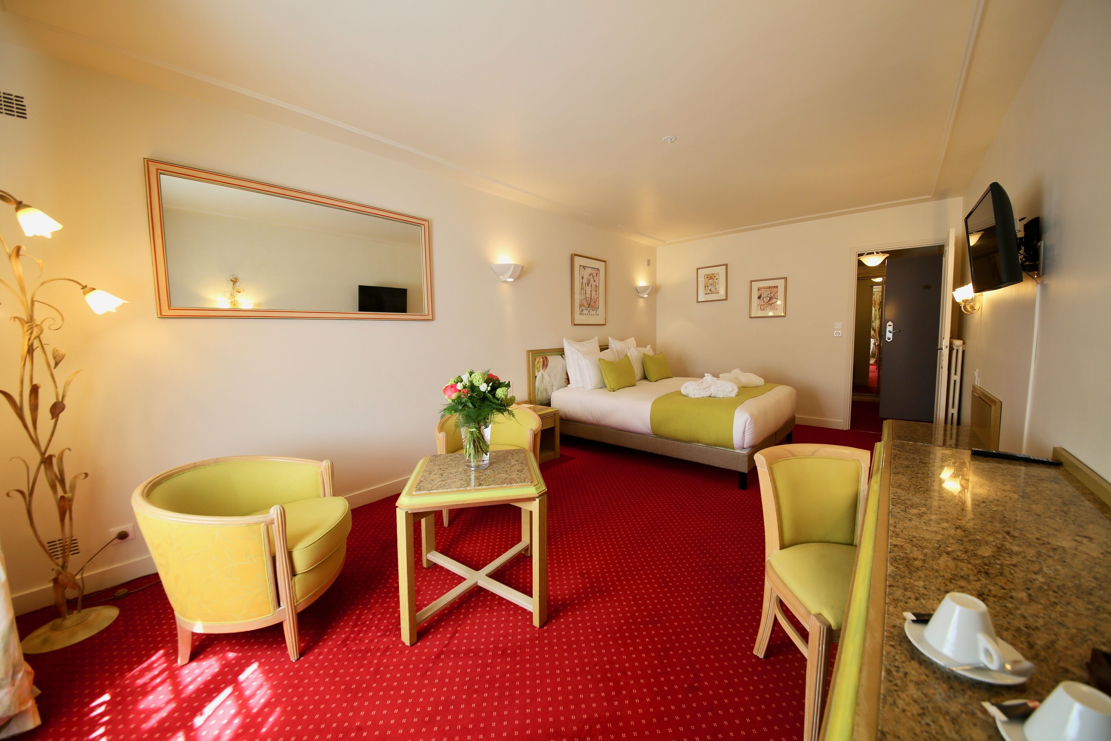 Habitación de hotel con alfombra roja, cama con ropa de cama verde y blanca, sillón amarillo, mesita con un jarrón de flores y un espejo grande en la pared.