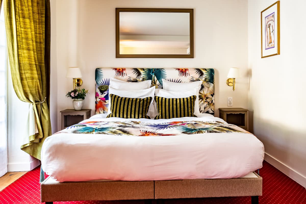 Habitación de hotel con una cama grande, dos mesitas de noche con lámparas, un espejo sobre la cama y un cuadro en la pared.