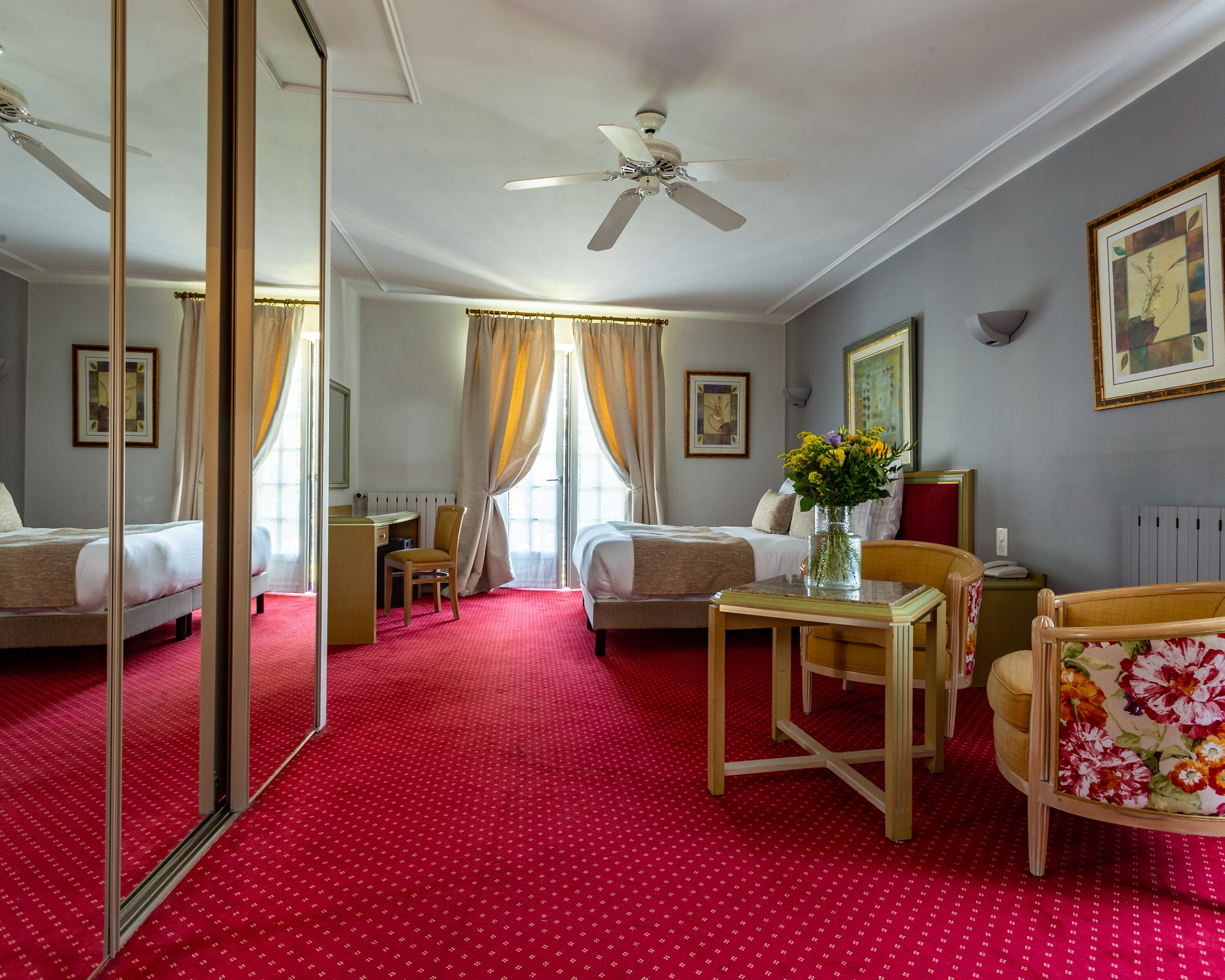 A hotel room with two beds, a desk, a chair, a table with a vase of flowers, and a ceiling fan.