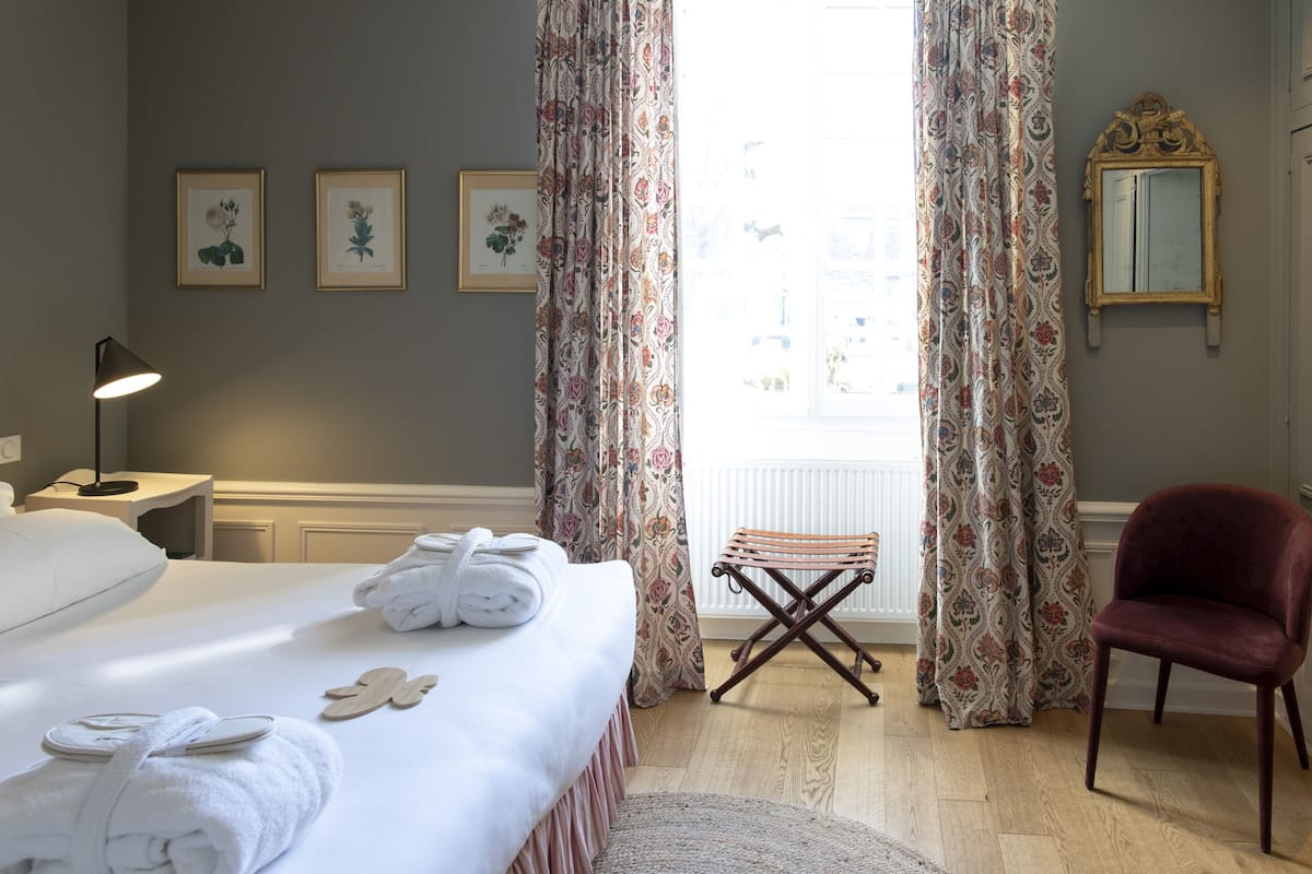 A neatly made bed with towels, a wooden bench, a red chair, and framed botanical prints on the wall.