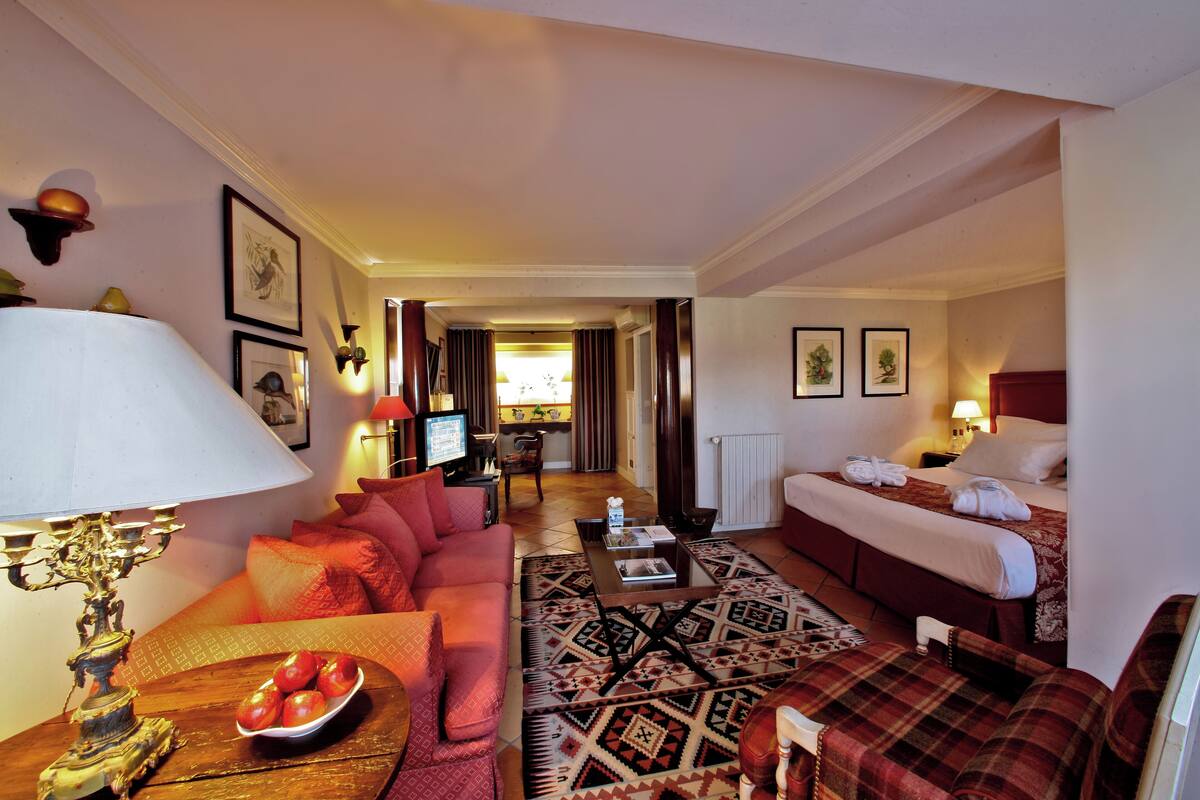 A hotel room with a sofa, a bed, a dining table with fruit, a patterned rug, and a lamp.
