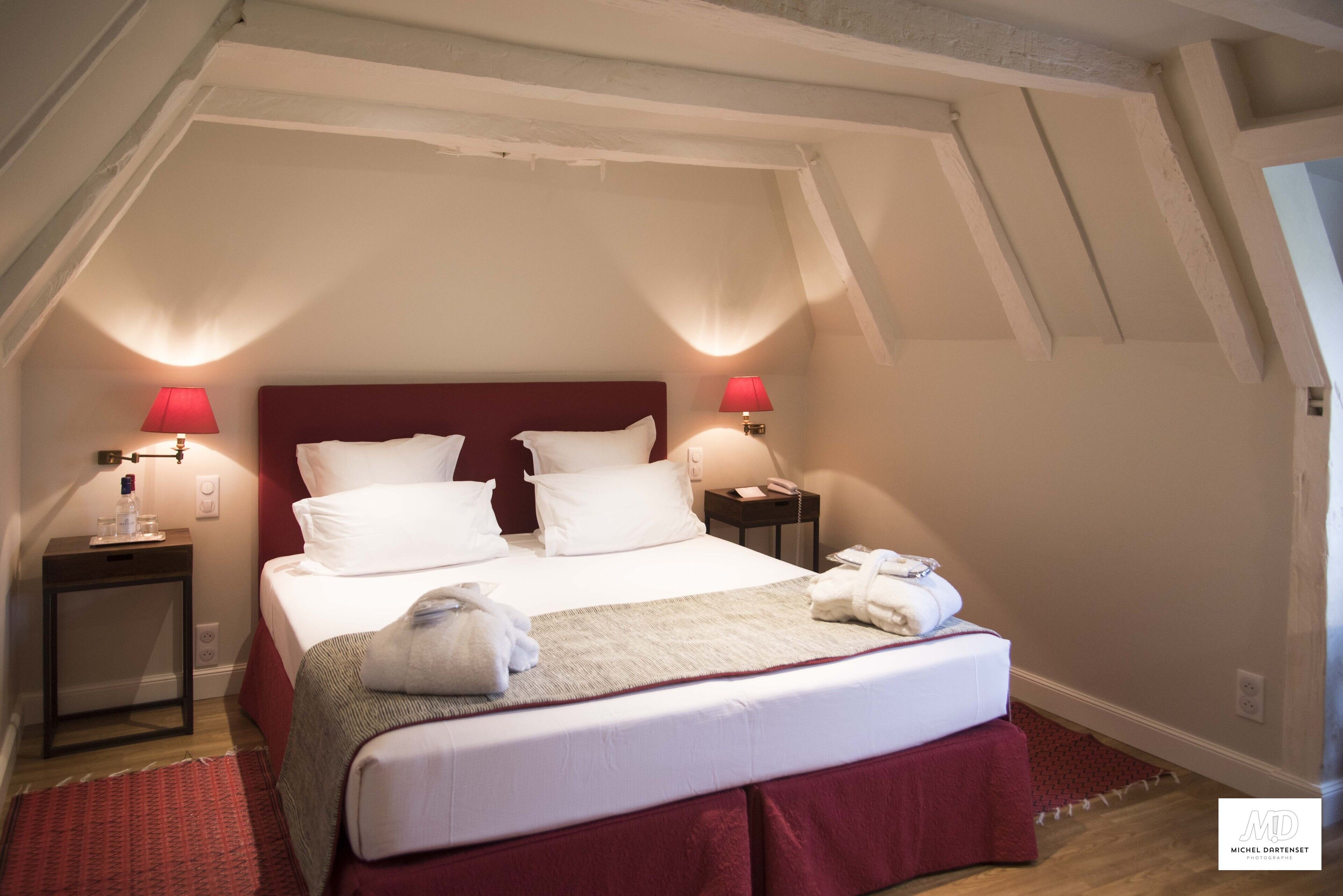 A neatly made bed with white linens and a red headboard in a room with sloped ceilings and two bedside lamps.