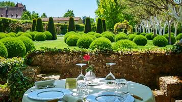 Un espacio de comedor al aire libre con una mesa para dos, rodeado por un jardín cuidado con setos recortados y un muro de piedra.