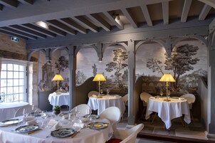 Uma sala de jantar com mesas preparadas para dois, cada uma com uma lâmpada, pratos e talheres. O espaço apresenta tetos em arco e um mural nas paredes.