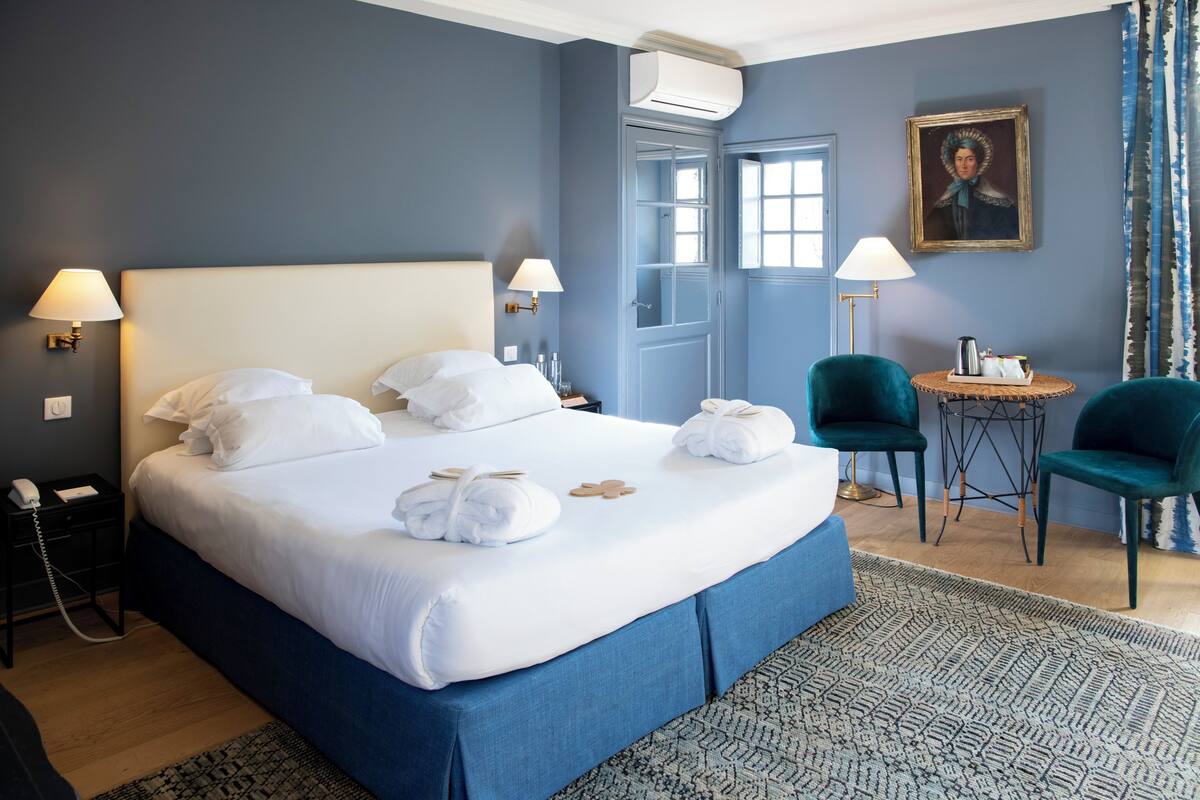 A hotel room with a large bed, two bedside lamps, a small table with a lamp, two chairs, and a painting on the wall.
