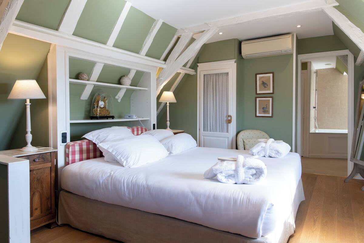 A neatly made bed with white linens and towels, a wooden nightstand with a lamp, a checkered pillow, and a shelf with decorative items.