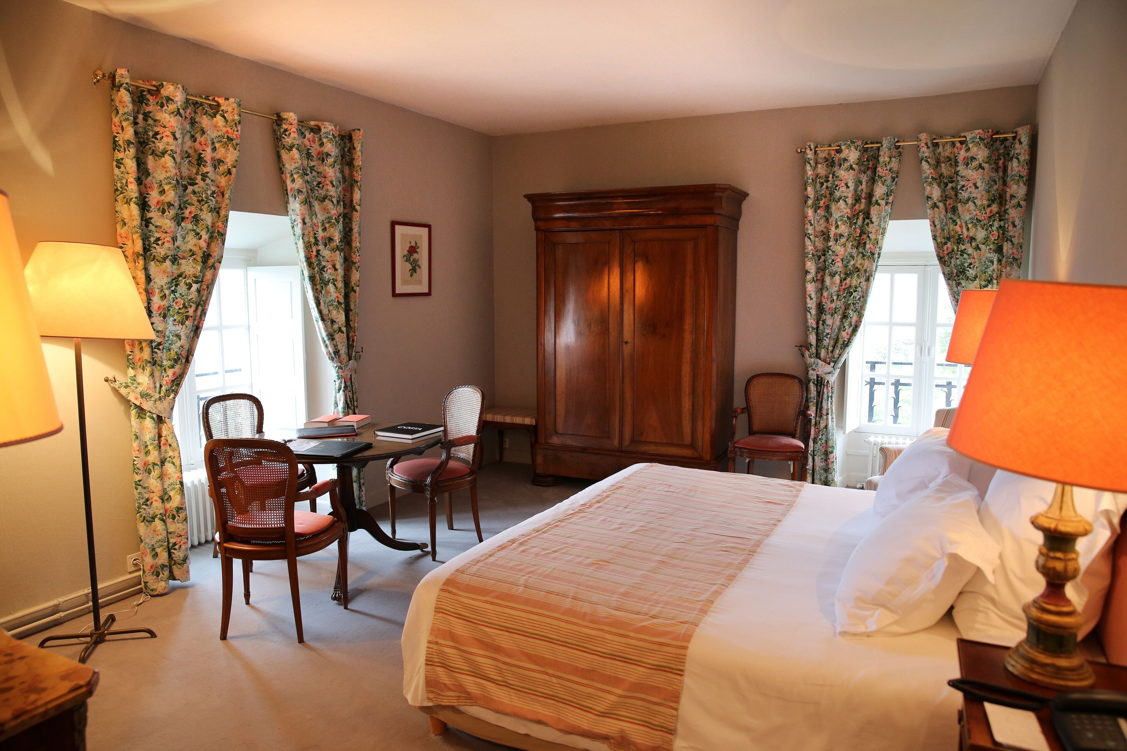 A hotel room with a bed, two chairs, a wooden wardrobe, and a table with a lamp.