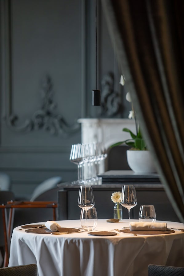 A dining table set with wine glasses, plates, and cutlery, with a vase of flowers and a potted plant in the background.