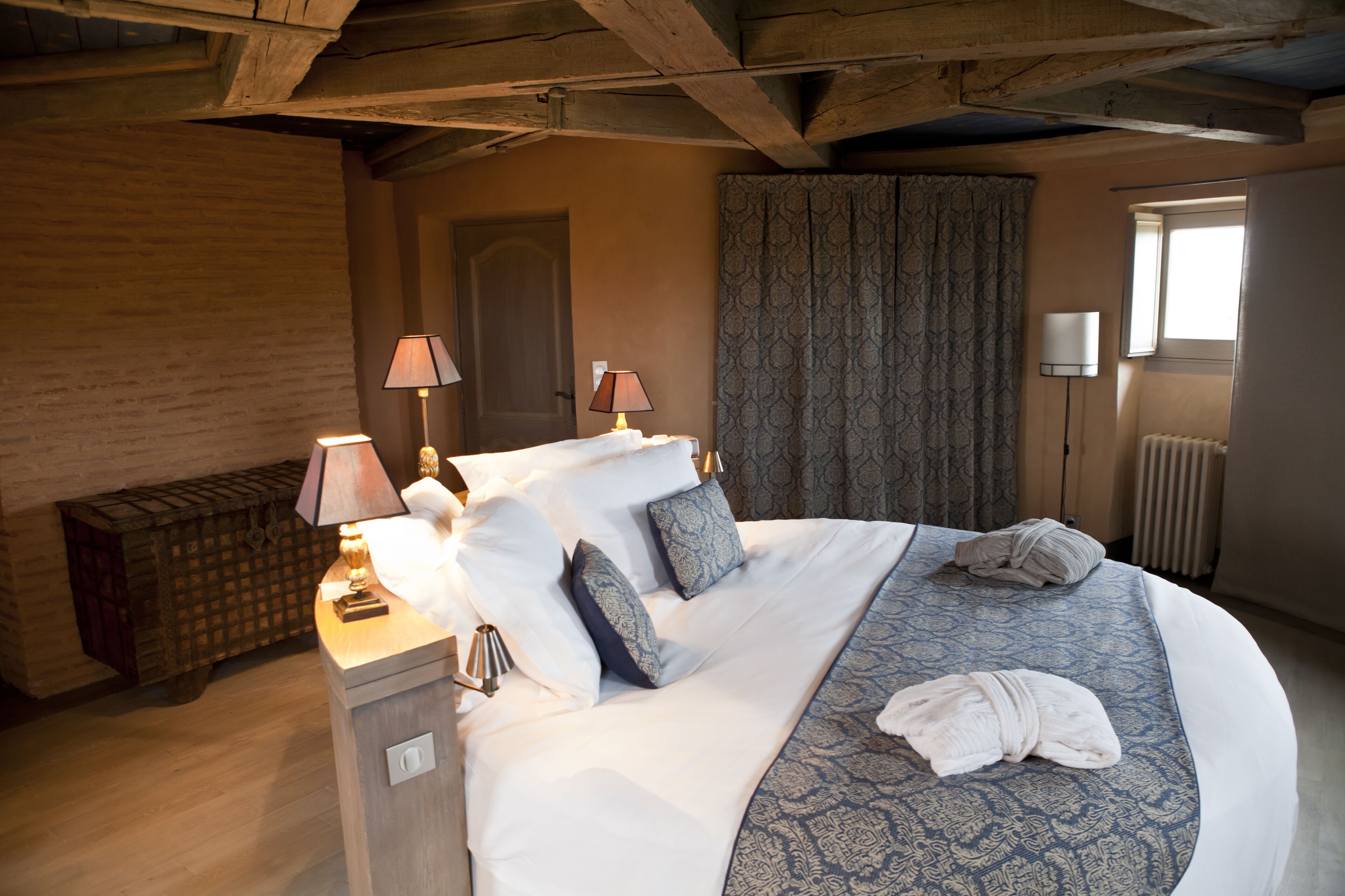 Une chambre d’hôtel avec un grand lit, deux lampes de chevet, un banc en bois et une fenêtre avec des rideaux.
