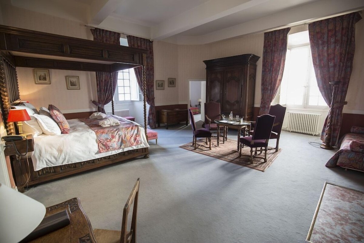 A traditional bedroom with a four-poster bed, a sitting area with a table and chairs, and a large window with curtains.
