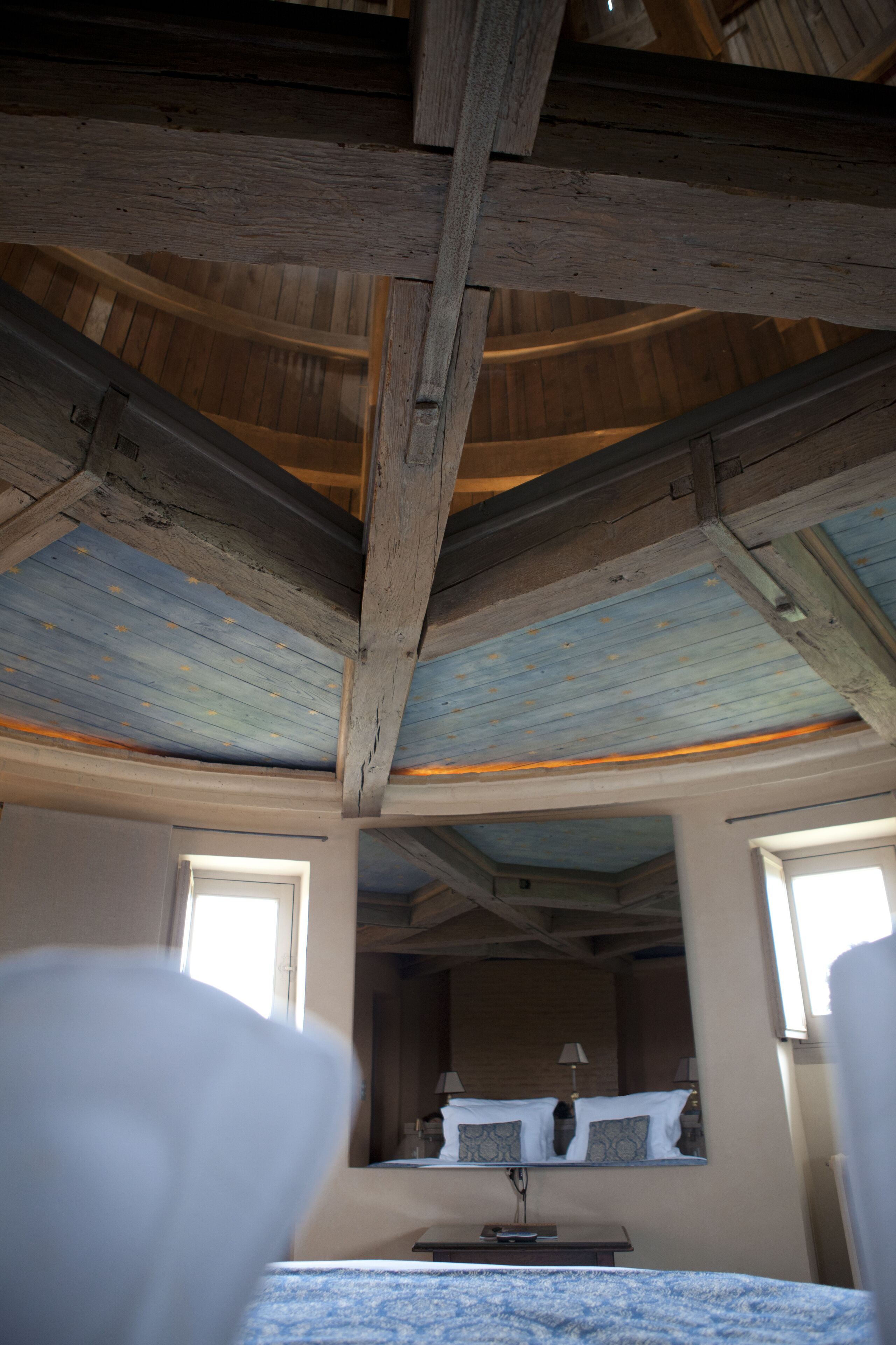 Una habitación con vigas de madera a la vista, una cama con manta azul, una mesita de noche y un espejo que refleja la habitación.