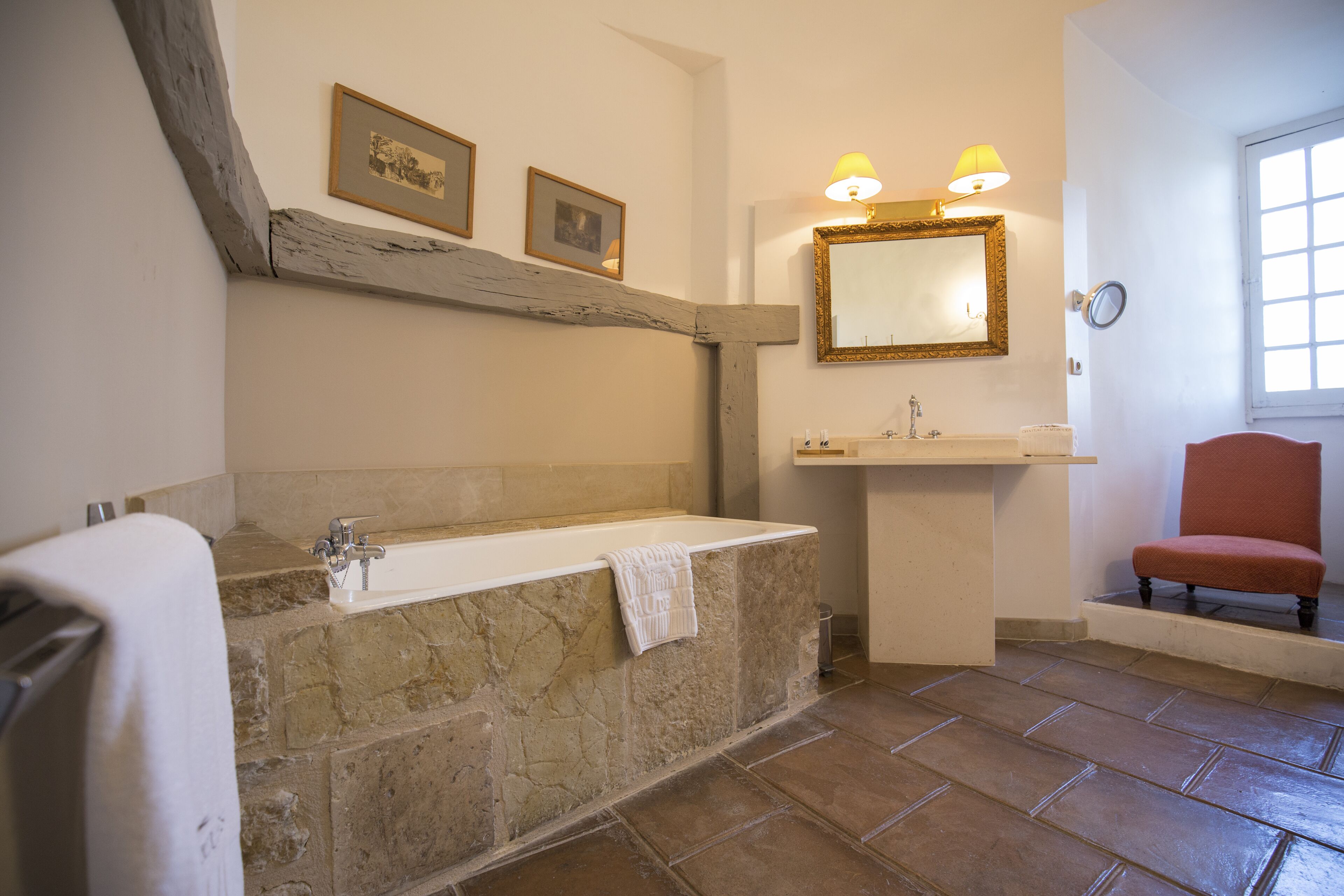 A bathroom with a stone bathtub, a sink, a mirror with a gold frame, and a red chair.