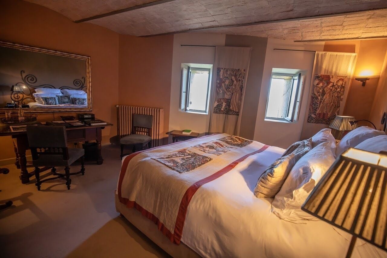 Un dormitorio con una cama grande, un escritorio, una silla y un espejo. Hay dos ventanas con cortinas, un radiador y una lámpara de pared.