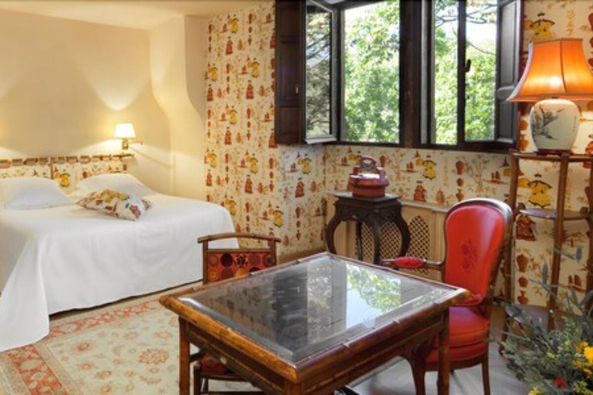 A room with two beds, a glass-top table, a red chair, and a window with a view of greenery.